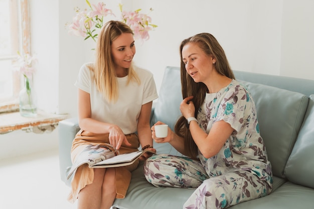 Zwei entzückende Frauen unterhalten sich zu Hause auf dem Sofa, trinken Kaffee und schauen sich ein Modemagazin an