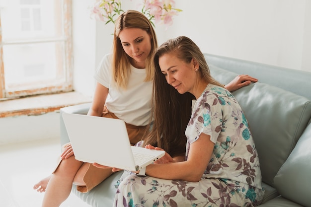 Zwei entzückende junge Frauen unterhalten sich zu Hause auf der Couch, trinken Kaffee und arbeiten an einem Laptop