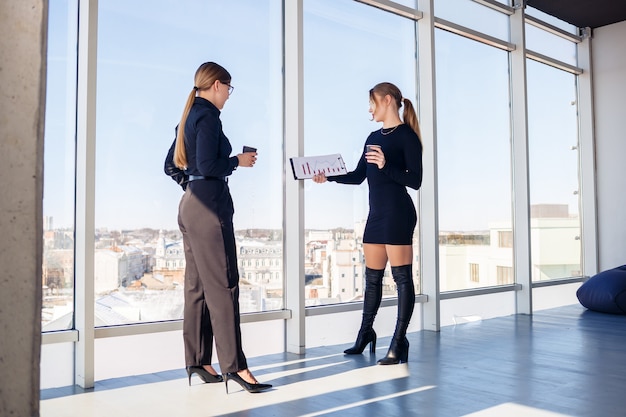 Zwei erfolgreiche Managerinnen unterhalten sich am Fenster im Büro über ein gemeinsames Projekt. Junge Ökonominnen in festlicher Kleidung unterhalten sich in ihren Pausen und trinken Kaffee.