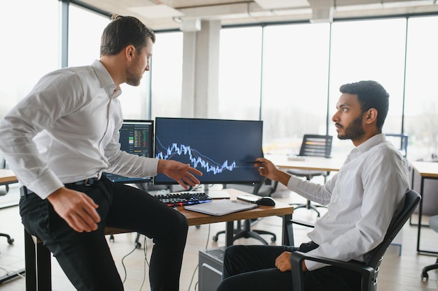 Zwei erfolgreiche Trader sitzen im Büro und schauen sich den Bildschirm an, um die Daten der Kryptowährungsinformationen auf dem Finanzmarktdiagramm zu überprüfen, das auf den Monitor zeigt