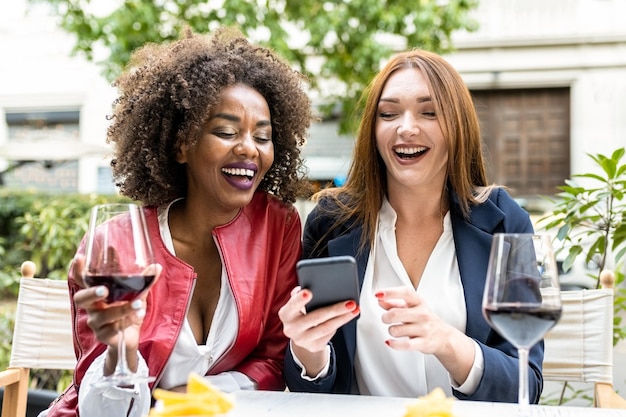 Zwei erwachsene Frauen, die sich während der Happy Hour nach der Arbeit amüsieren, lachen, während sie sich das soziale Netzwerk des Smartphones und die mobile App für techniksüchtige Menschen ansehen