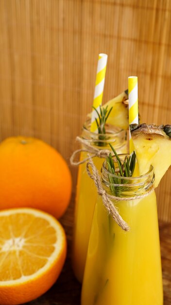 Zwei Flaschen tropischer Saft mit Papierstrohhalmen. Orangen, Ananas und Rosmarin zur Dekoration. Hölzerner Hintergrund. Vertikales Foto