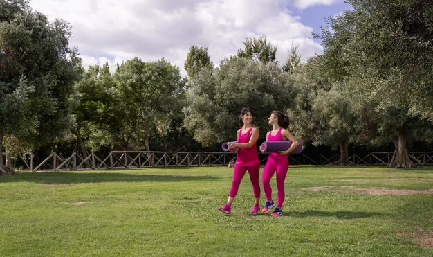 Zwei Frauen, die an einem sonnigen Tag mit gerollten Pilated-Matten in einem Park spazieren gehen. Junge und kaukasische Frauen mittleren Alters in rosa Kleidung im Freien. Sport und gesundes Lifestyle-Konzept