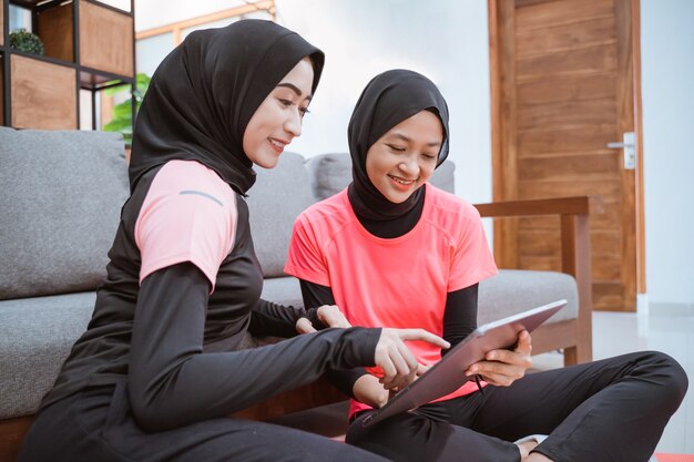 Zwei Frauen, die Hijab-Sportkleidung tragen, während sie entspannt auf dem Boden sitzen, während sie ein digitales Tablet verwenden und den Tablet-Bildschirm mit ihren Fingern berühren, wenn sie sich zu Hause auf das Sofa lehnen