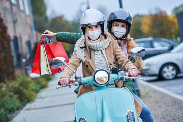 zwei Frauen, die Masken tragen und Einkaufstüten halten, die auf dem Roller pendeln