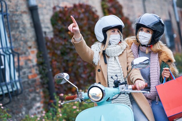 zwei Frauen, die Masken tragen und Einkaufstüten halten, die auf dem Roller pendeln