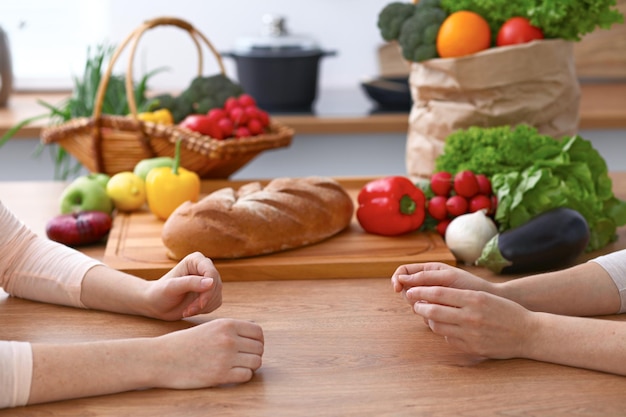 Zwei Frauen diskutieren in der Küche über ein neues Menü, Nahaufnahme. Menschliche Hände von zwei Personen, die zwischen frischem Gemüse am Tisch gestikulieren. Koch- und Freundschaftskonzept.