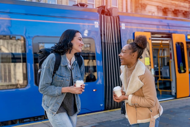 Zwei Frauen in einer Jeansjacke unterhalten sich lachend, trinken Kaffee und warten auf eine Straßenbahn