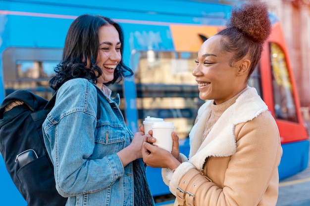 Zwei Frauen in einer Jeansjacke unterhalten sich, trinken Kaffee und warten an der Haltestelle Lifestyle photo auf die Straßenbahn