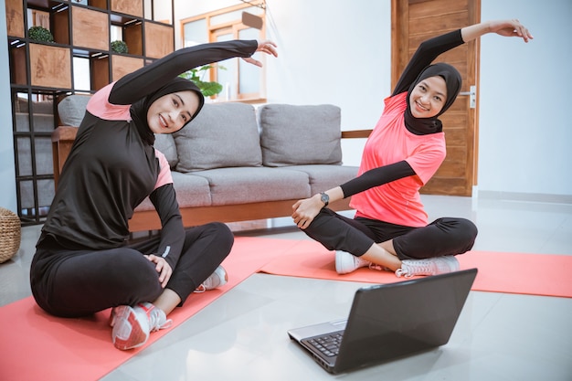 Zwei Frauen in Hijab-Sportbekleidung sitzen mit gekreuzten Beinen auf dem Boden. Ihre Körper sind zur Seite gelehnt und die Hände hoch, während sie gemeinsam ihre Arme im Haus aufwärmen