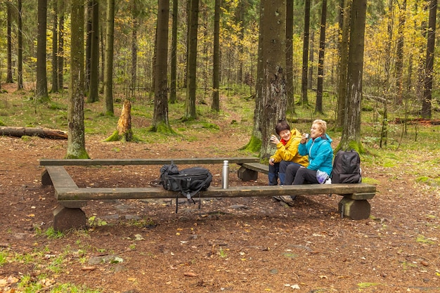 Zwei Frauen lachen und sitzen auf einer Bank im Wald. Trinken Sie Tee oder Kaffee aus einer Thermoskanne