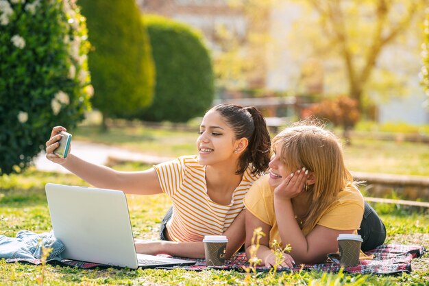 Zwei Frauen Latina im Park liegend auf einer Decke mit Computer und Kaffee, die an einem sonnigen Tag ein Selfie machen