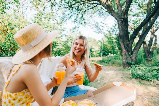Zwei Frauen sitzen auf einem Sofa im Hinterhof und essen Pizza, trinken Limonade an einem sonnigen Sommertag.