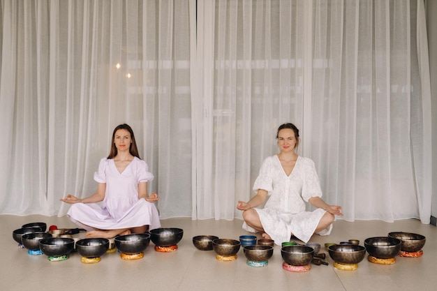 Zwei Frauen sitzen mit tibetischen Schalen im Lotussitz vor einer Yogastunde im Fitnessstudio