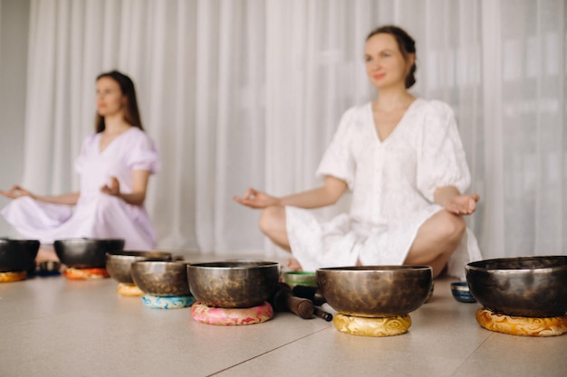 Zwei Frauen sitzen mit tibetischen Schalen im Lotussitz vor einer Yogastunde im Fitnessstudio