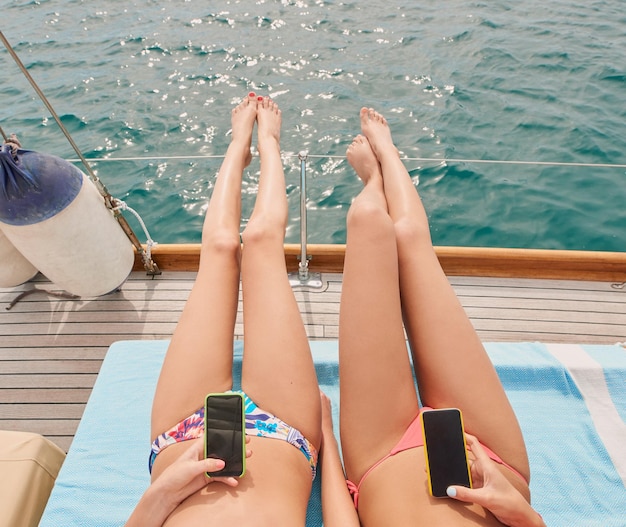 Zwei Freunde beim Sonnenbaden auf dem Boot während der Urlaubskreuzfahrt zusammen mit Handys, um Textnachrichten zu senden Zwei Frauen, die mit Smartphones auf dem Boot liegen und sich in Bikinis sonnen