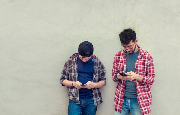 Zwei Freunde im Teenageralter an einer Wand, die ihre Handys überprüft Zwei Freunde, die sich an eine Wand lehnen, die auf ihren Telefonen SMS schreibt Freund, der seinem Freund Handy zeigt Lächelnde Freunde, die Handys überprüfen