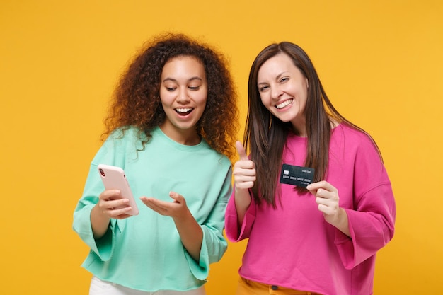 Zwei freundinnen europäisch-amerikanische mädchen in rosa grüner kleidung posieren isoliert auf gelbem hintergrund. People-Lifestyle-Konzept. Mock-up-Kopienbereich. Halten Sie Mobiltelefon, Kreditkarte und zeigen Sie den Daumen nach oben.