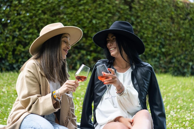 Zwei Freundinnen in Hüten, die auf dem Gras sitzen und mit einem Glas Wein rösten Lebensstil der jungen Freunde in Hüten, die auf dem Gras sitzen und mit Wein rösten