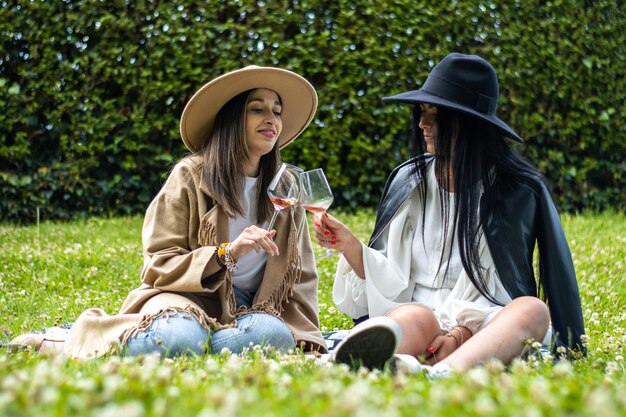 Zwei Freundinnen in Hüten, die auf dem Gras sitzen und mit einem Glas Wein rösten Lebensstil der jungen Freunde in Hüten, die auf dem Gras sitzen und mit Wein rösten