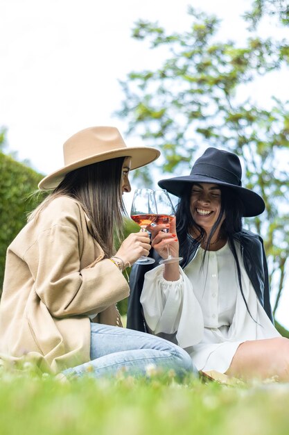 Zwei Freundinnen in Hüten, die auf dem Gras sitzen und mit einem Glas Wein rösten Lebensstil der jungen Freunde in Hüten, die auf dem Gras sitzen und mit Wein rösten