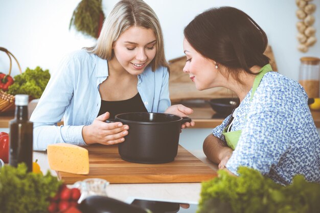 Zwei Freundinnen schauen mit einem Fertiggericht in den dunklen Topf und probieren neue Rezepte, während sie am Küchentisch sitzen Gesundes Kochkonzept