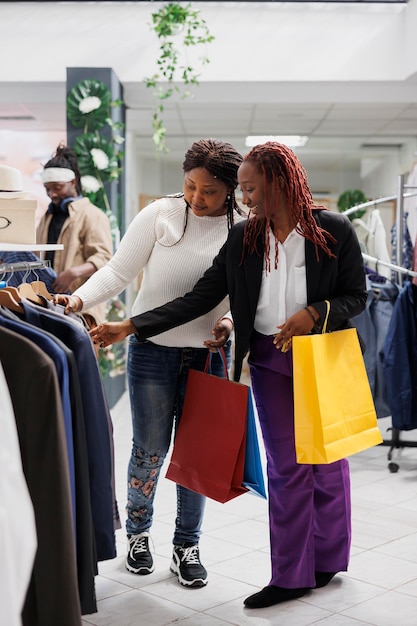 Zwei Freundinnen tragen Einkaufstüten und stöbern im Jackenregal im Einkaufszentrum. Fröhliche afroamerikanische Frauen mit Papierpaketen, die sich in einer Modeboutique für einen Blazer entscheiden