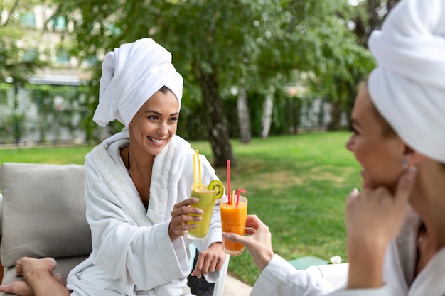 Zwei freundliche Frauen in weißen Bademänteln, die mit Getränken im Spa sitzen und sich nach der Spa-Pflege miteinander unterhalten