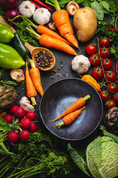 Zwei frische Bauernkarotten in Vintage-Schüssel und verschiedenes Bio-Gemüse auf rustikalem schwarzem Betonhintergrund. Herbsternte, vegetarisches Essen oder sauberes gesundes Essenskonzept kochen, Draufsicht