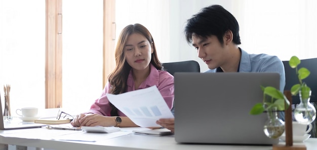 Zwei fröhliche asiatische junge Geschäftsleute, die mit einem Tablet in einem modernen Büro zusammenarbeiten
