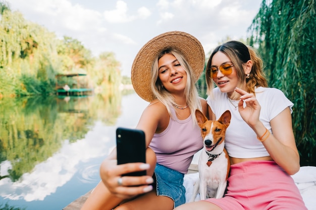 Zwei fröhliche Freundinnen ruhen sich im Freien auf dem Seepier mit Hund aus, machen Foto, Selfie