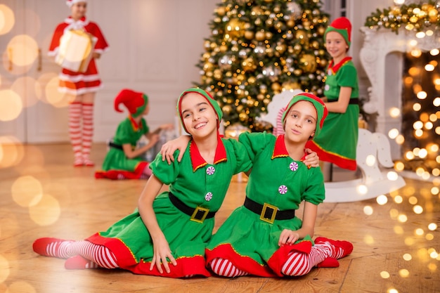 Zwei fröhliche, fröhliche Freundinnen in Kostümen von Weihnachtselfen auf dem Hintergrund eines Weihnachtsbaums und anderer Kinder