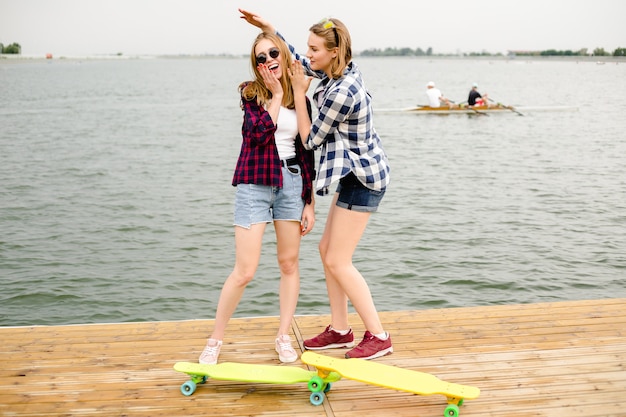 Zwei fröhliche, glückliche Skatermädchen im Hipster-Outfit, die sich während der Sommerferien auf einem Holzsteg amüsieren.