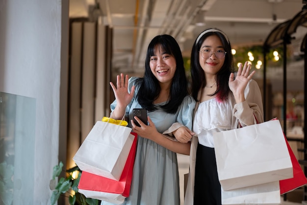 Zwei fröhliche junge asiatische Frauen winken glücklich in die Kamera, während sie Einkaufstaschen in der Hand halten