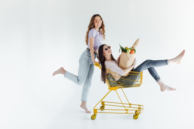 Zwei fröhliche junge Frauen in farbenfrohen trendigen Outfits lächeln und haben Spaß mit Trolley