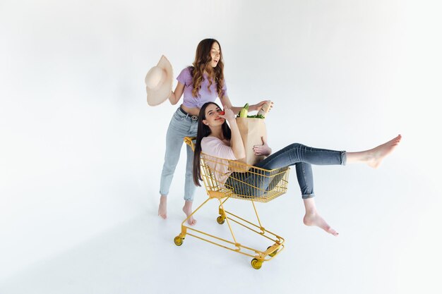 Zwei fröhliche junge Frauen in farbenfrohen trendigen Outfits lächeln und haben Spaß mit Trolley