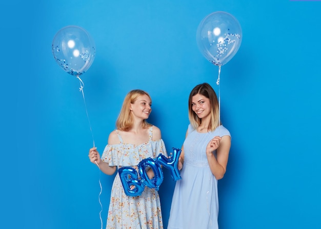 Zwei fröhliche junge kaukasische Modelle, die blaue Luftballons und einen Ballon mit dem blauen Hintergrund des Aufschriftjungen halten