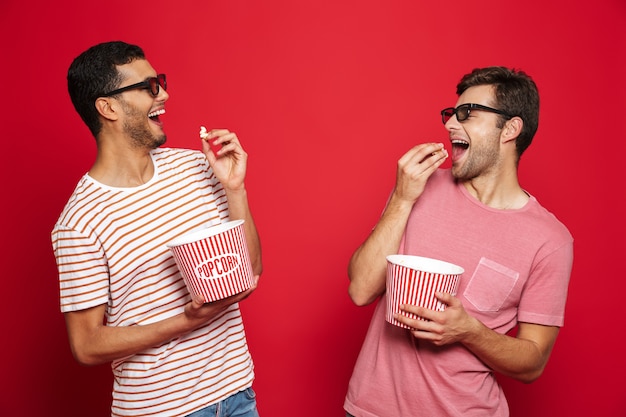 Zwei fröhliche junge Männer, die isoliert über roter Wand stehen, Popcorn essen, 3D-Brille tragend