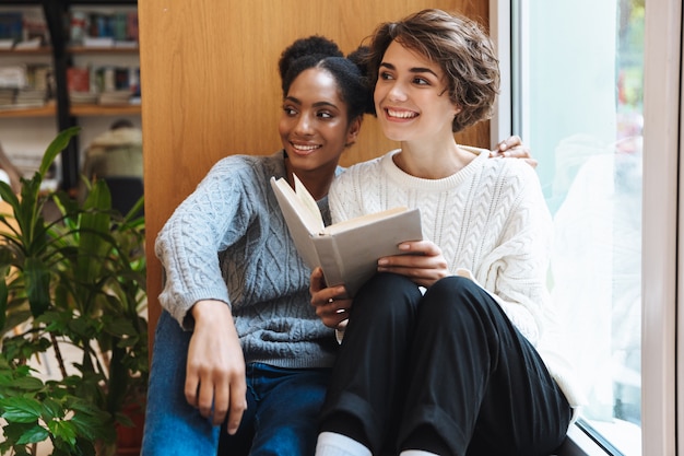 Zwei fröhliche junge Studentinnen, die in der Bibliothek studieren und gemeinsam ein Buch lesen