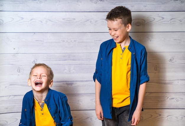 Zwei fröhliche Jungen in blauen Hemden und gelben T-Shirts lachen zusammen im Studio Fröhliche Brüder genießen die Zeit und haben gemeinsam Spaß isoliert auf grauem Holzhintergrund