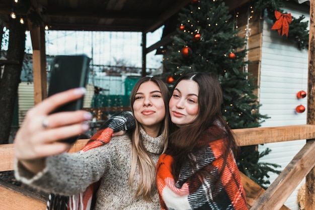 Zwei fröhliche kaukasische Freundinnen, die Spaß haben und Selfie auf dem Hinterhof mit Weihnachtsdekorationen machen.