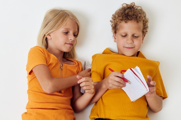 Zwei fröhliche Kinder liegen mit Notizblöcken und Bleistiften auf dem Boden, isolierter Hintergrund unverändert