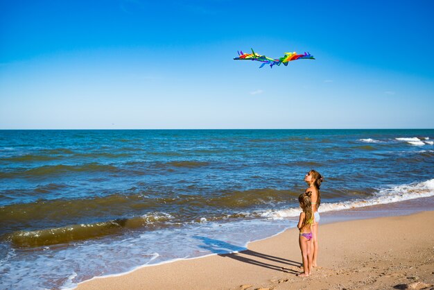 Zwei fröhliche kleine Mädchenschwestern spielen mit einem Drachen, der an einem sonnigen warmen Sommertag am sandigen Ufer am Meer läuft