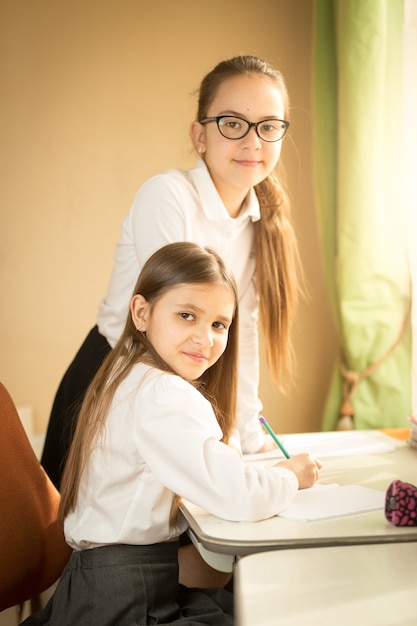 Zwei fröhliche Mädchen in Schuluniform posieren hinter Schreibtisch