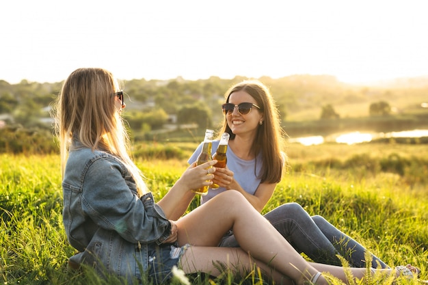 Zwei fröhliche Mädchen und junge Freunde mit Sonnenbrille, Bier trinken und gemeinsam die Zeit bei Sonnenuntergang genießen.