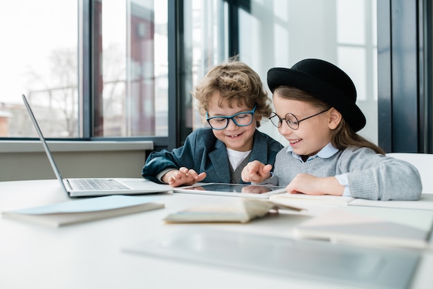 Zwei fröhliche Schulfreunde mit digitalem Tablet, die durch Online-Informationen schauen, während sie am Schreibtisch sitzen