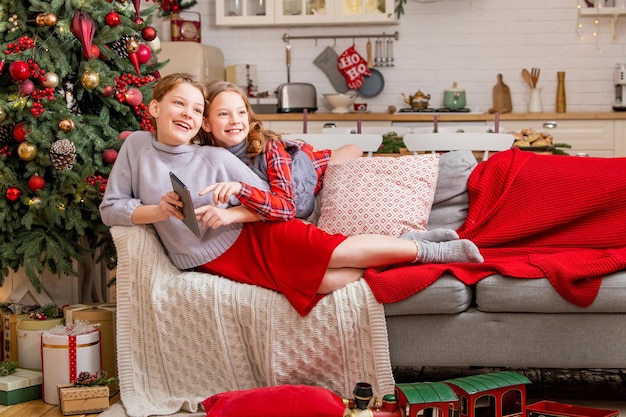 Zwei fröhliche Schwestern sitzen zu Hause neben dem Weihnachtsbaum und schauen auf den Tablet-Monitor