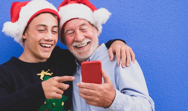 Zwei fröhliche Weihnachtsmänner, die sich an einer blauen Wand umarmen, ein Großvater mit seinem Enkel im Teenageralter, lachen, während sie zusammen auf das Handy schauen. Konzept der modernen und technologischen Familie