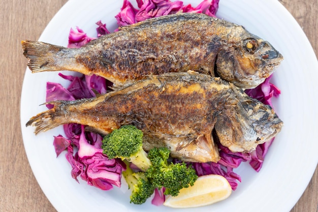 Zwei gegrillte Meeresfische, serviert auf einem Teller mit Rotkohl, grünem Brokkoli und Zitrone Nahaufnahme