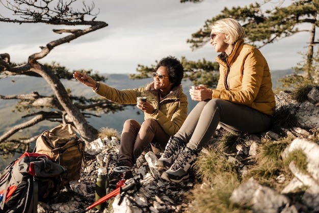 Zwei gemischtrassige Freundinnen sitzen auf einem Hügel und machen während ihrer Wanderung in den Bergen an einem schönen sonnigen Herbsttag eine Teepause.